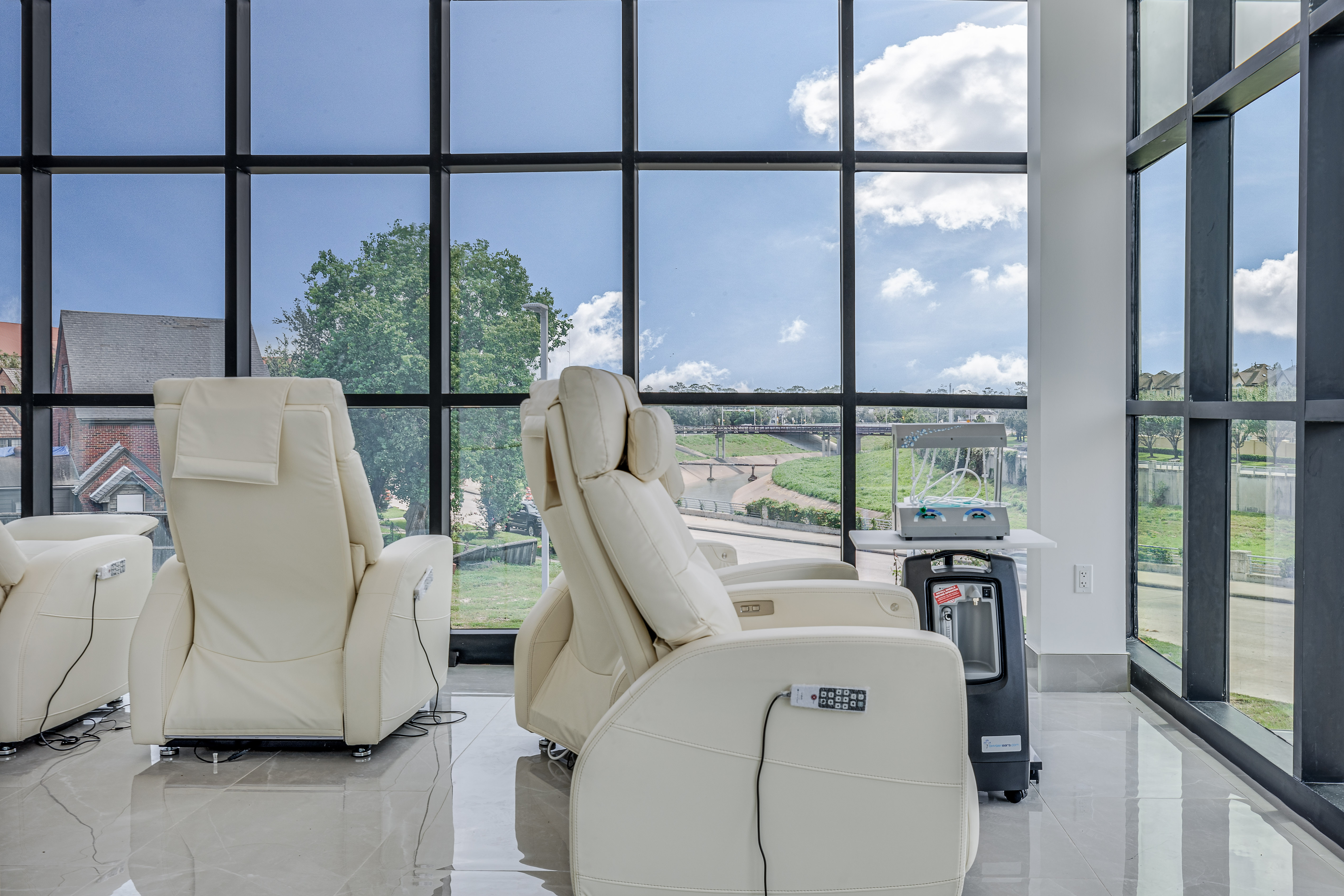 Cream leather reclining lounge chairs with oxygen concentrators beside floor-to-ceiling windows overlooking the bayou